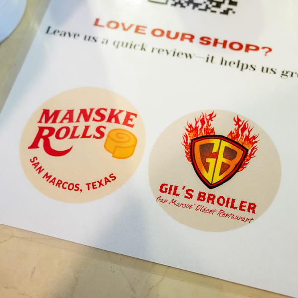 Two circular stickers for Manske Rolls and Gil's Broiler on a white paper with a review request.