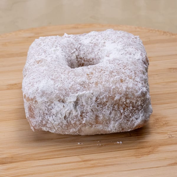 A square Manske Roll donut, covered in powdered sugar, rests on a light wooden surface.