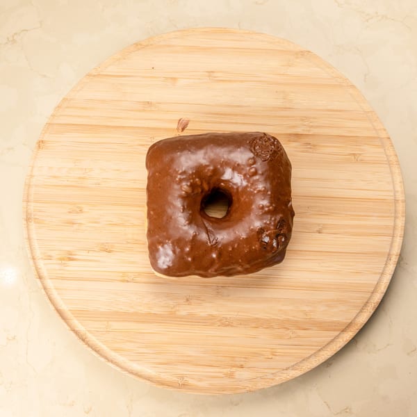 A square chocolate-glazed Manske Roll donut sits on a round wooden cutting board.