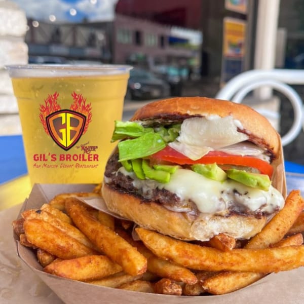 Gil's Broiler burger with avocado, cheese, lettuce, tomato, fries, and a drink on an outdoor table.